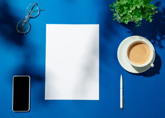 A blue table with a blank white piece of paper, a cell phone, a pair of glasses, a pen, and a cup of coffee on it